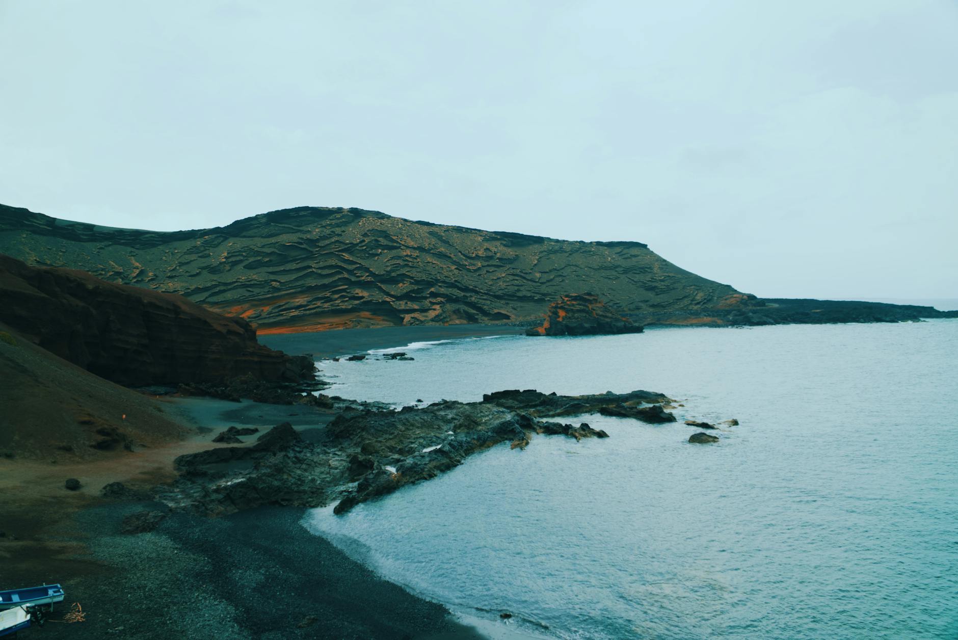 Eine felsige Kueste mit einem schwarzen Strand und dem Meer unter einem bewölkten Himmel.