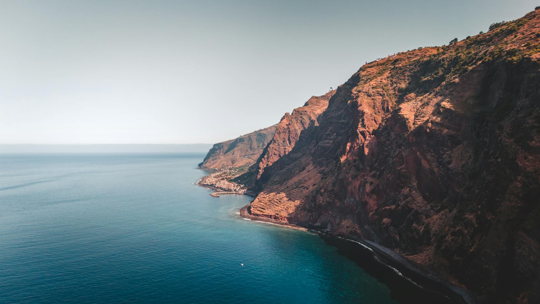Eine steile, felsige Küste mit einem kleinen Dorf und einem Hafen, der ins tiefblaue Meer ragt.