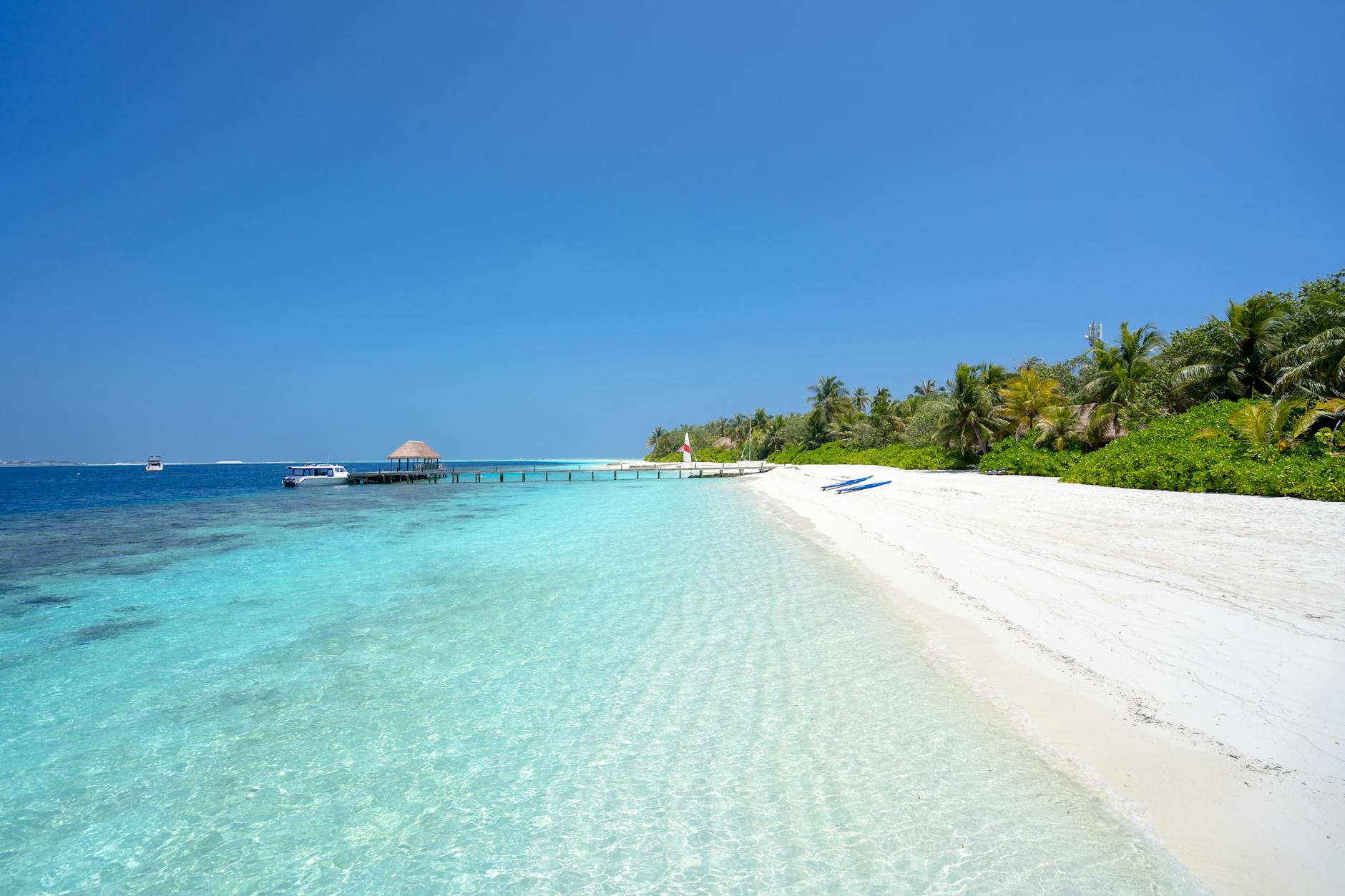 Ein weisser Sandstrand mit Palmen und gruener Vegetation, daneben ein Steg mit einem Boot im tuerkisen Wasser und ein klares blaues Meer.