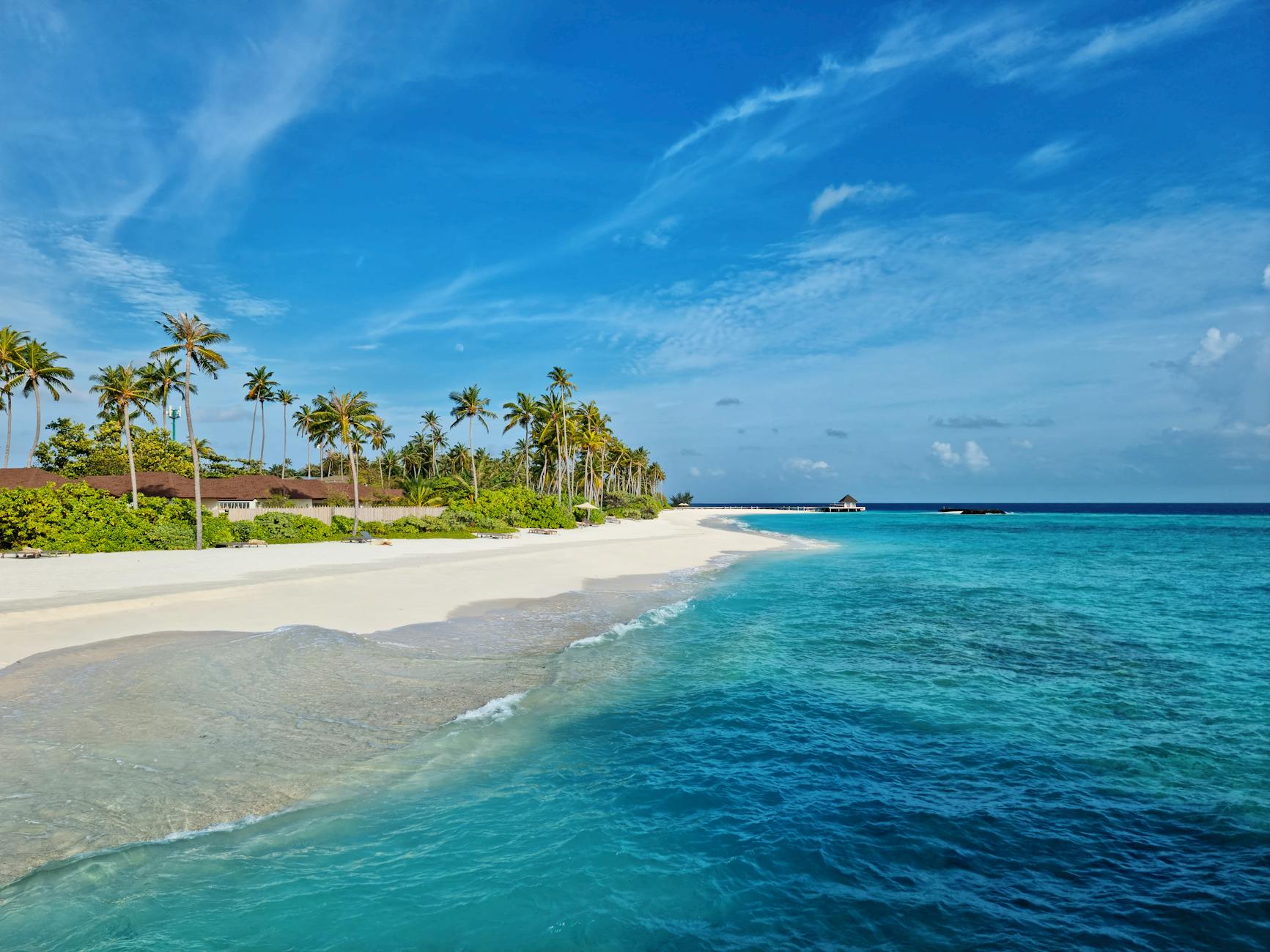 Ein weißer Sandstrand mit Palmen und türkisfarbenem Wasser, im Hintergrund eine Hotelanlage und ein Wasserbungalow.