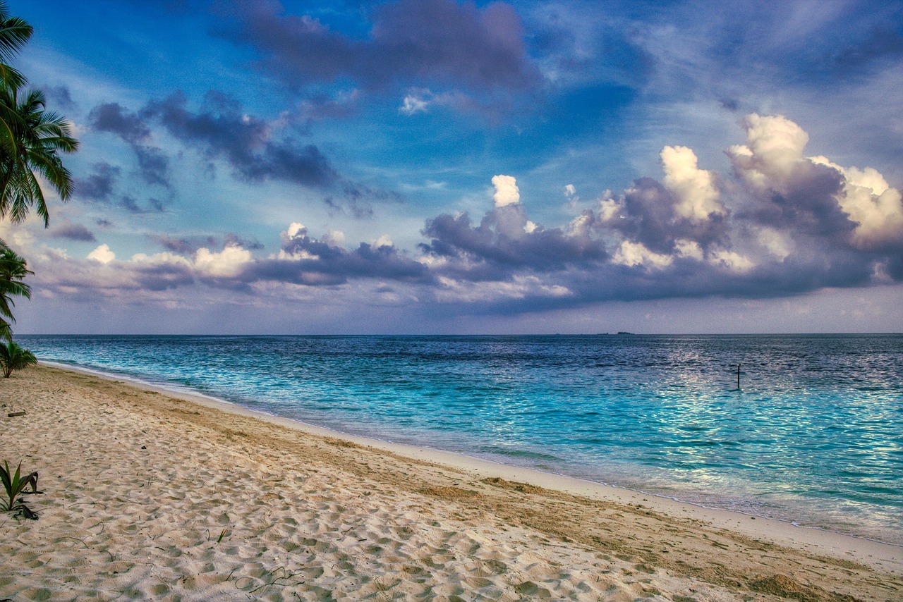 Ein Sandstrand mit Palmen am linken Bildrand und dem Meer mit tuerkisblauem Wasser und Wolken am Himmel.