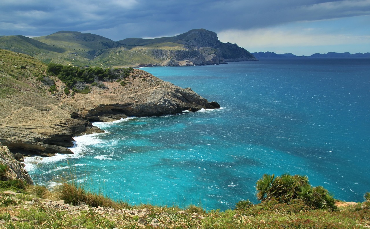 Eine felsige Küste mit türkisblauem Wasser und Wellen, die gegen die Felsen schlagen, im Hintergrund Berge.