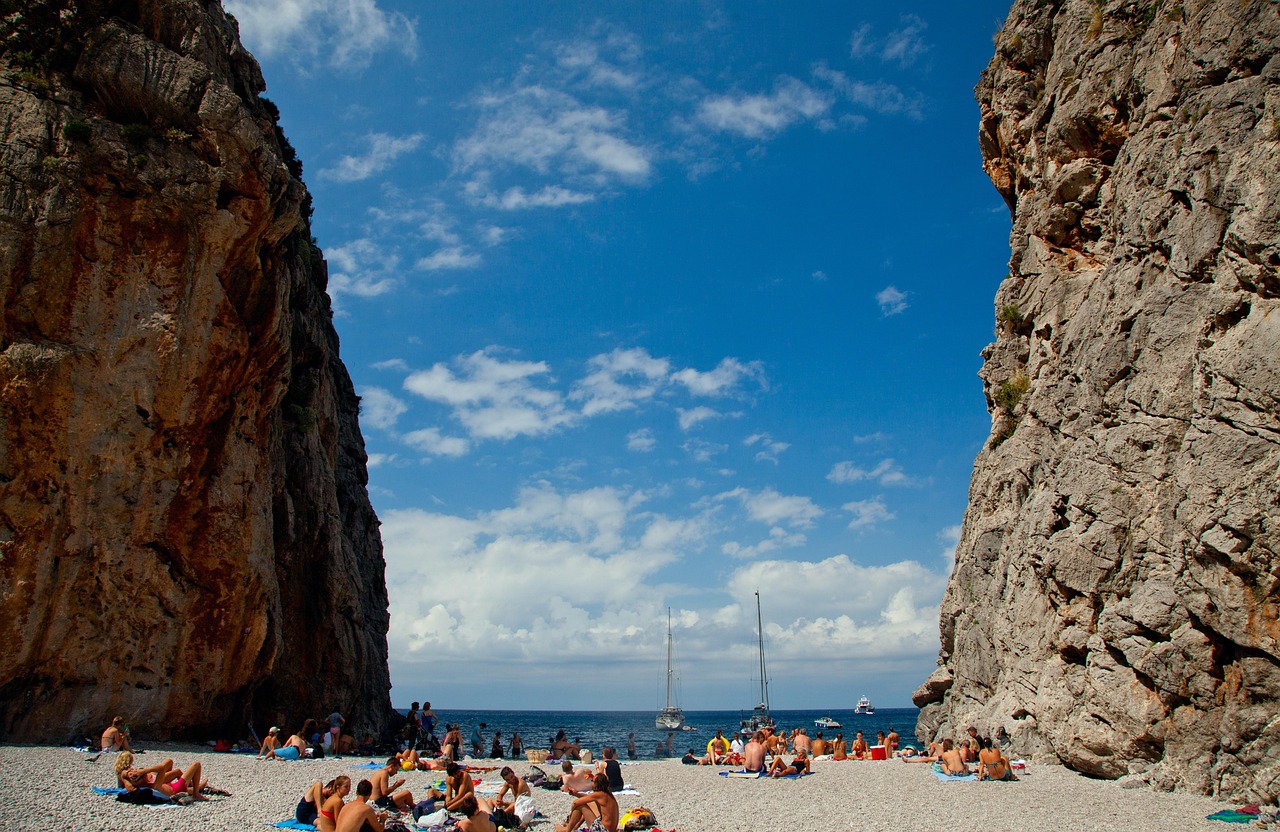 Ein Kiesstrand mit vielen Menschen, die auf Handtüchern liegen und sich sonnen, eingerahmt von zwei steilen Felswänden. Im Hintergrund ist das Meer mit einigen Booten zu sehen.