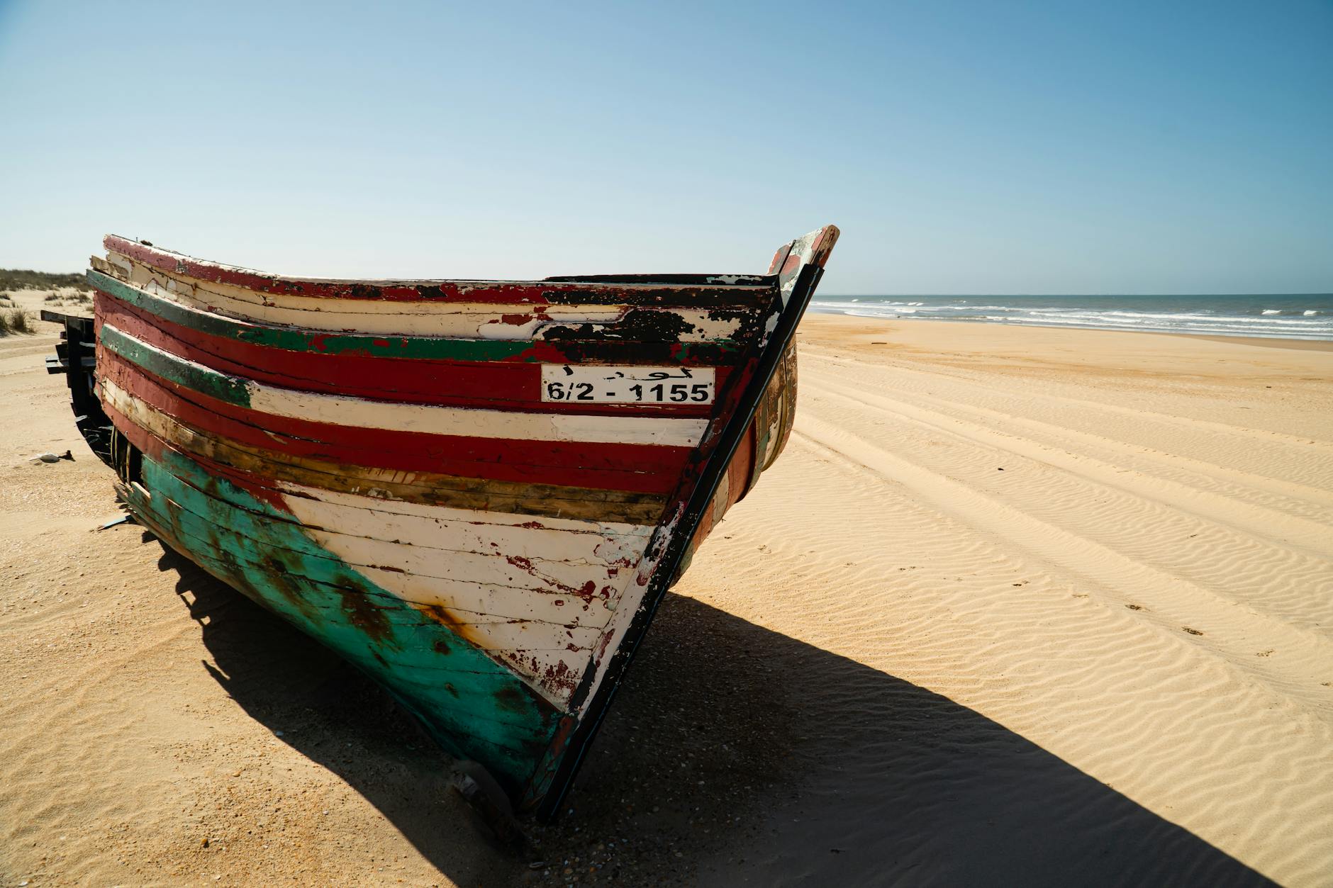 Ein bunt gestreiftes, altes Fischerboot liegt am sandigen Strand mit dem Meer im Hintergrund.