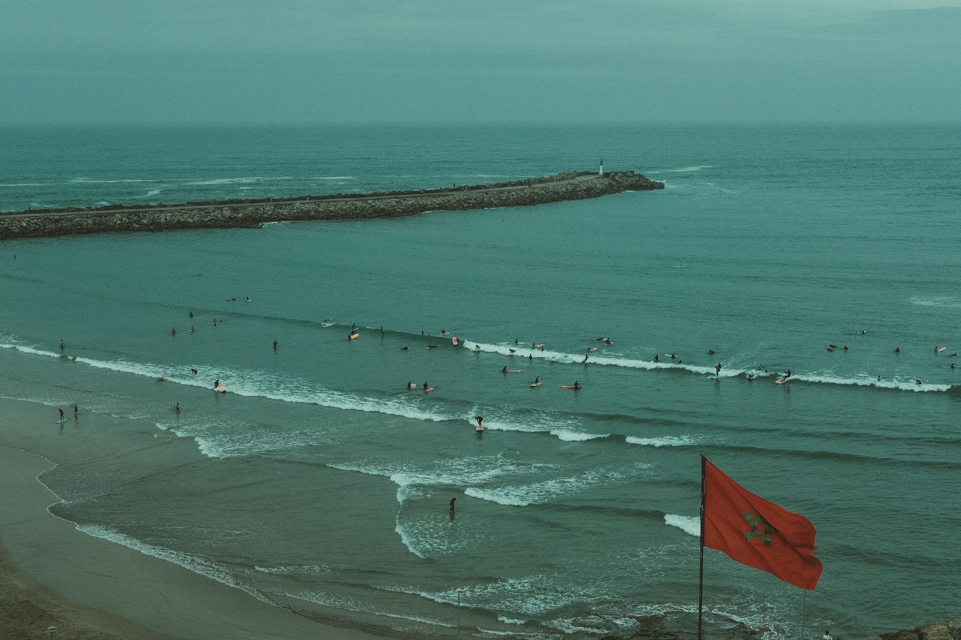 Eine marokkanische Flagge weht im Vordergrund vor einem Meer mit vielen Surfern und einem langen Pier mit einem Leuchtturm am Ende.