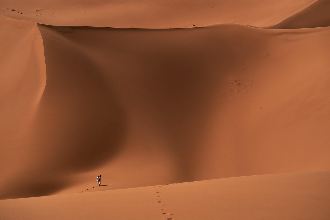 Eine Person wandert durch eine weite Dünenlandschaft in der Wüste und hinterlässt Spuren im Sand.