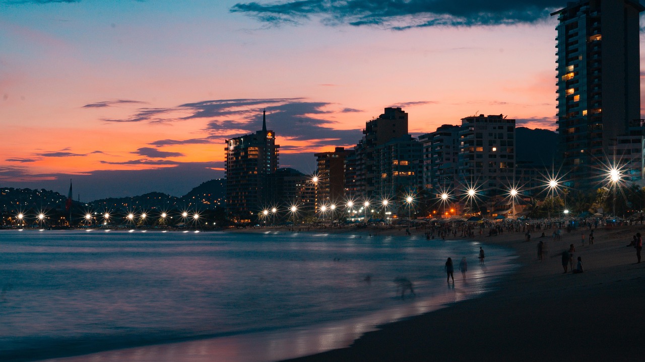 Eine Stadt am Meer bei Sonnenuntergang mit beleuchteten Gebaeuden und einem Strand mit Menschen.