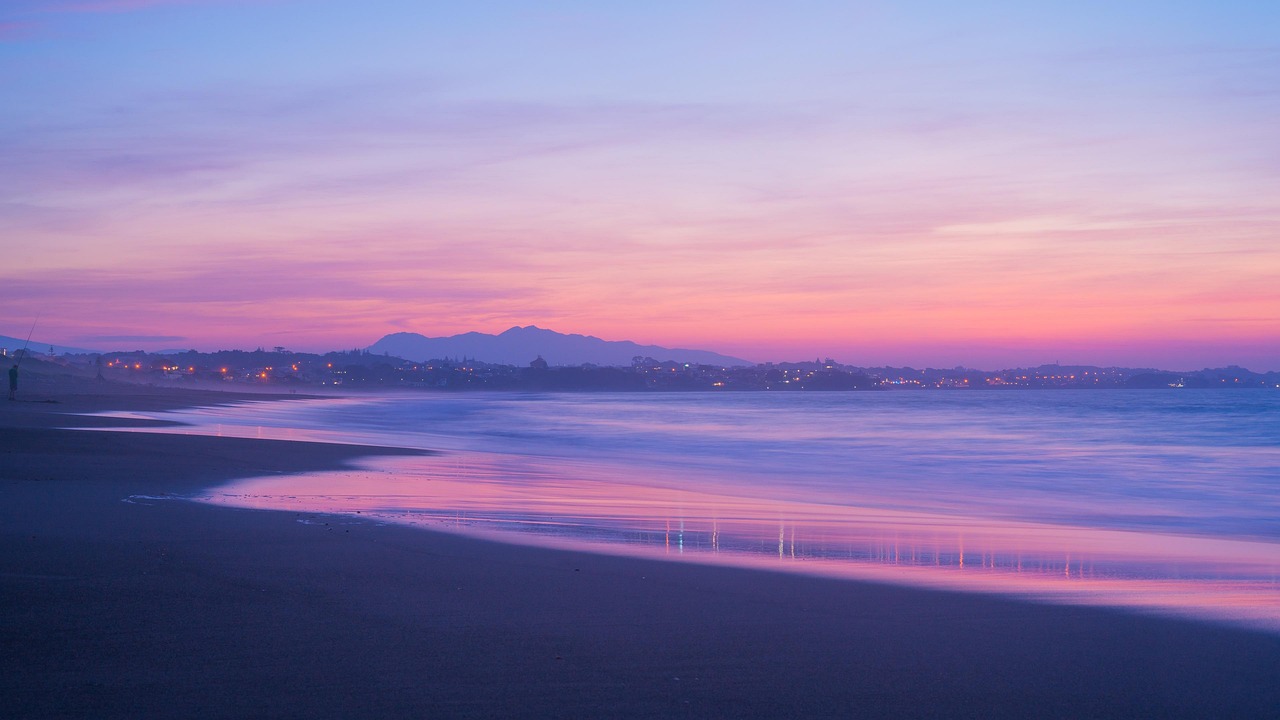 Ein Strand mit sanften Wellen und einer Stadt am Horizont bei Sonnenuntergang.