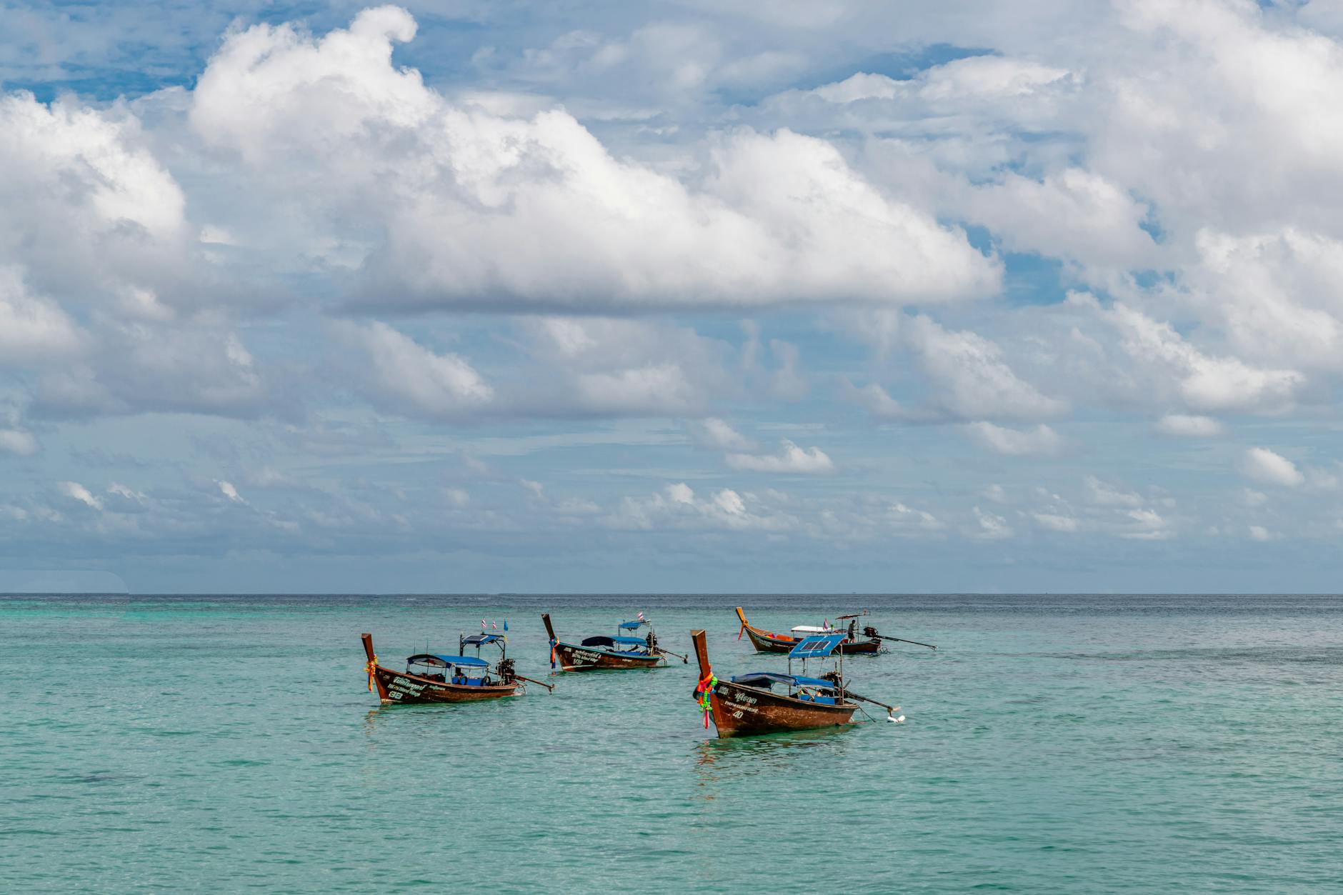 Mehrere Longtail-Boote treiben auf dem tuerkisen Meer unter einem Himmel mit Wolken.