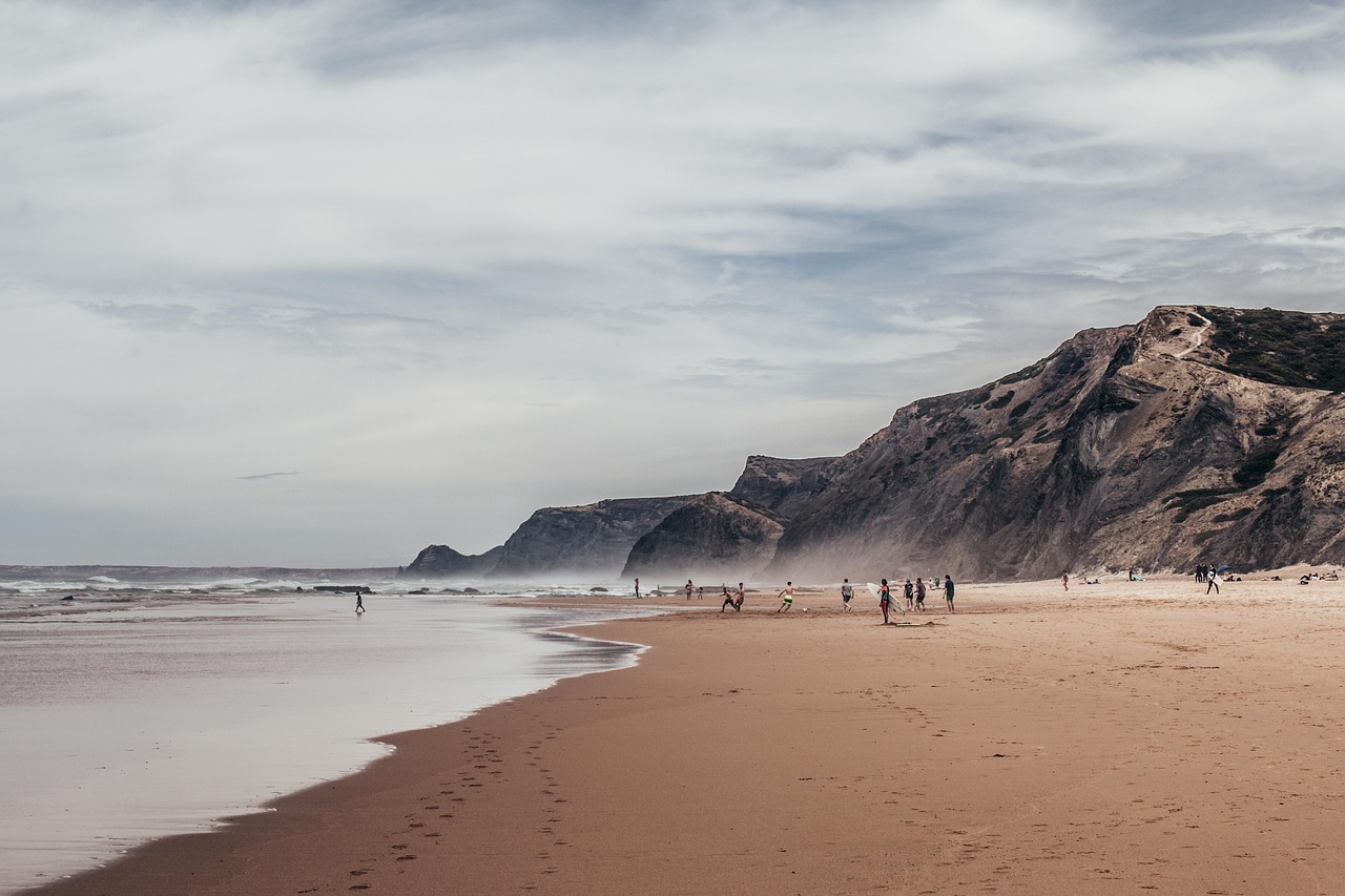Ein weiter Sandstrand mit Menschen, die am Ufer spielen, und steilen Felsformationen im Hintergrund unter einem bewölkten Himmel.