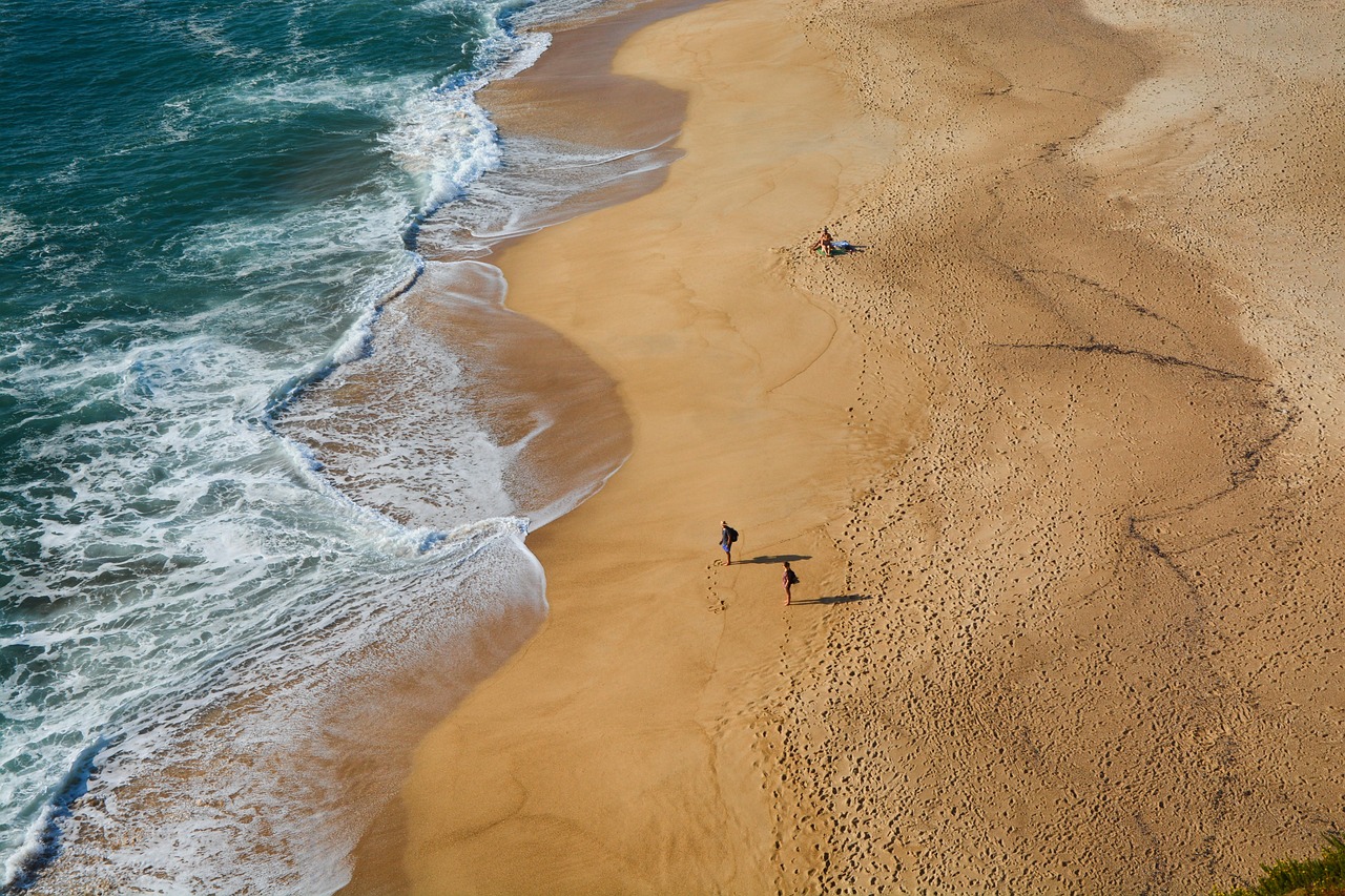 Luftaufnahme eines Sandstrandes mit Wellen, die auf den Strand treffen und wenigen Menschen, die spazieren gehen.