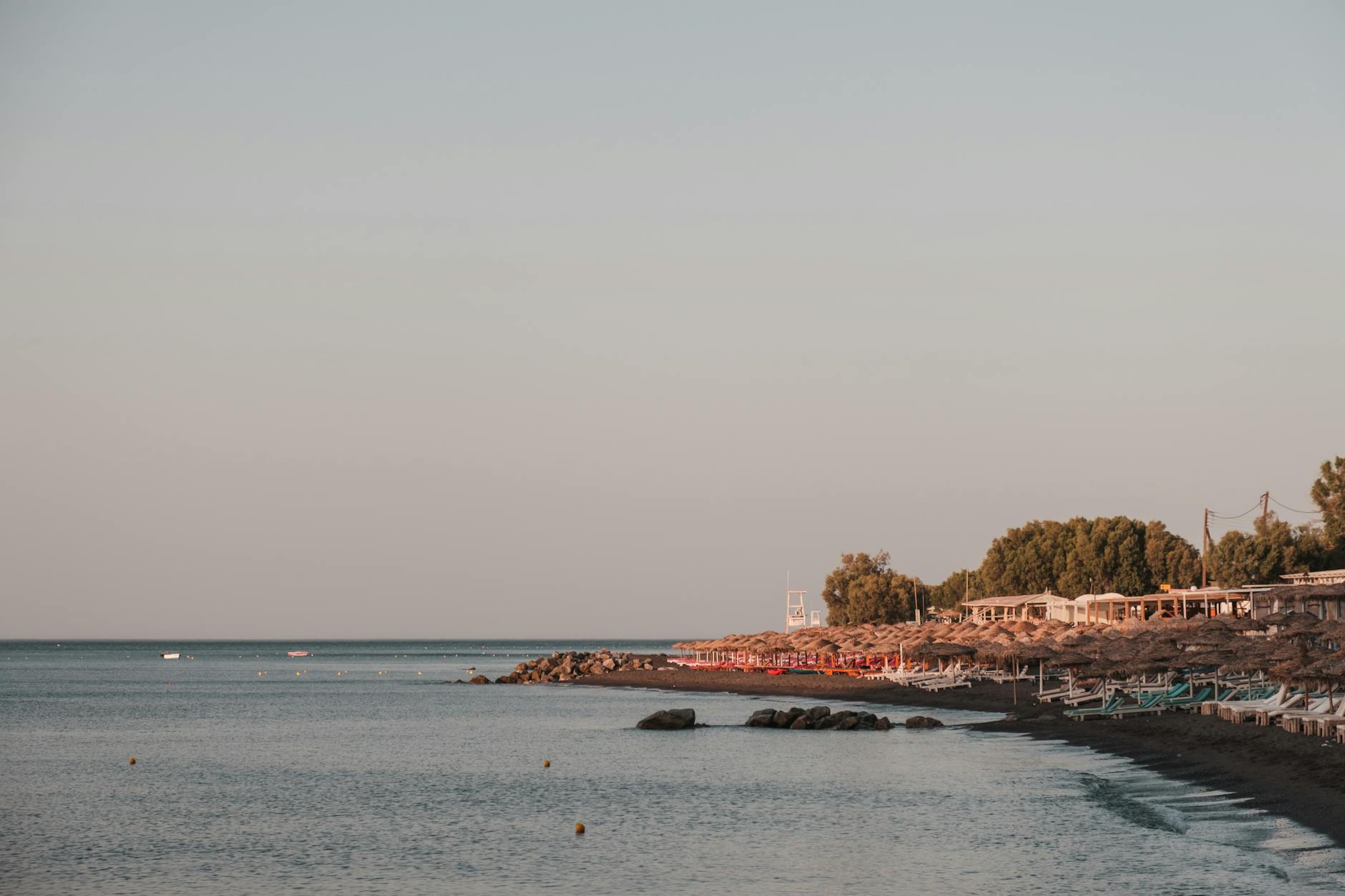 Ein Strand mit vielen Sonnenliegen und Strohschirmen, dahinter eine Reihe von Gebäuden und Bäumen, am Ufer des Meeres.