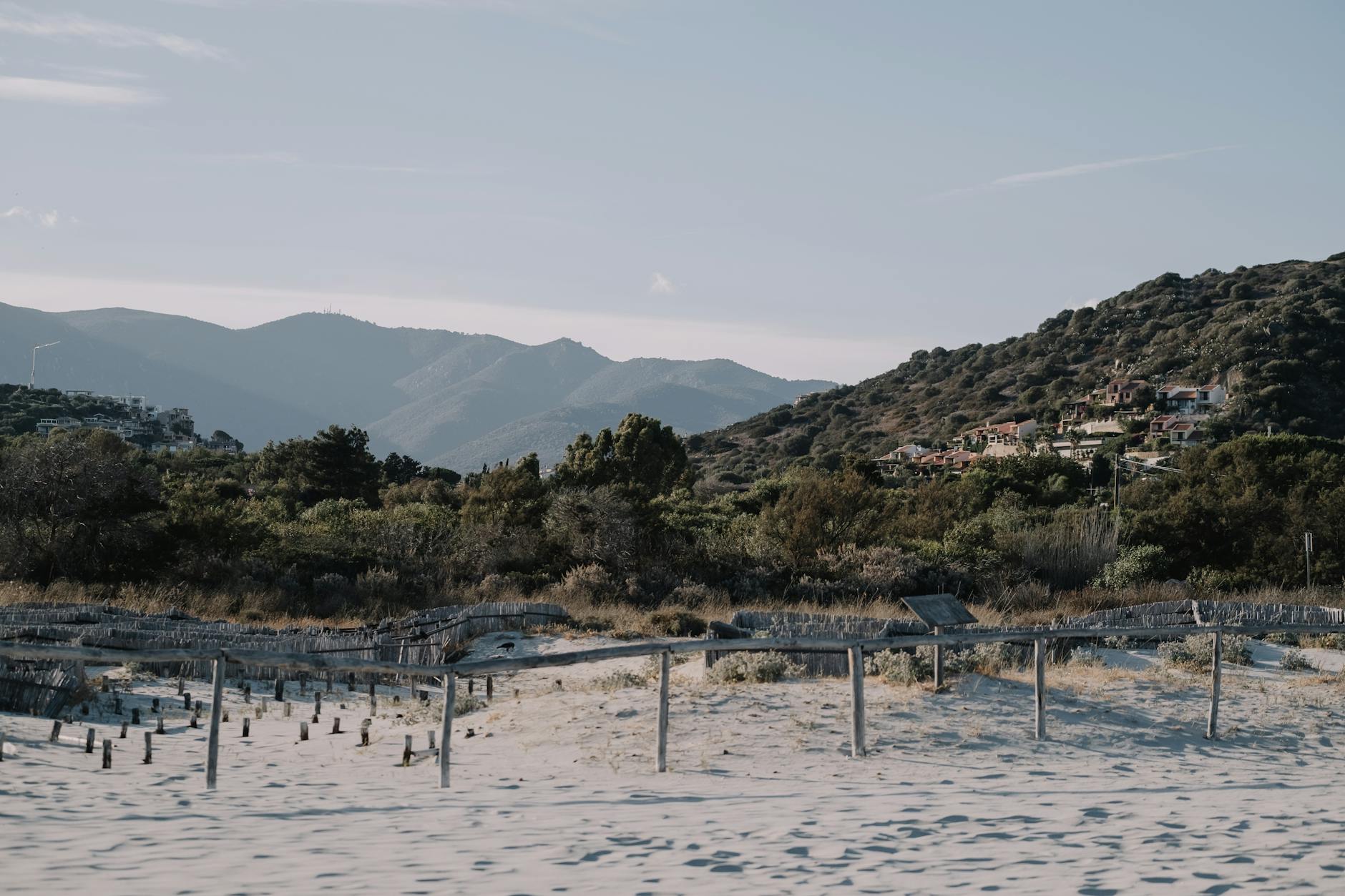 Ein sandiger Strand mit einem niedrigen Holzzaun und dahinterliegenden Dünen und mediterraner Vegetation, im Hintergrund erheben sich bewaldete Hügel mit vereinzelten Häusern.