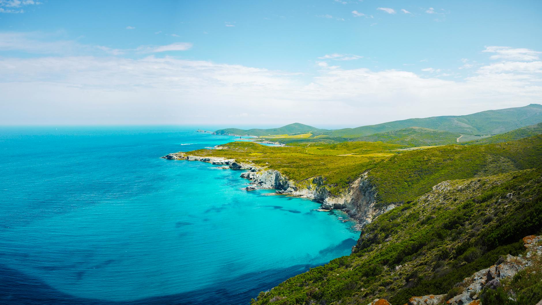 Blick auf eine felsige Kueste mit tuerkisblauem Wasser und gruenen Huegeln im Hintergrund unter einem blauen Himmel mit Wolken.