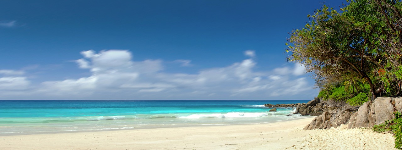 Ein einsamer Sandstrand mit tuerkisfarbenem Wasser und felsigen Klippen, bewachsen mit tropischer Vegetation, unter einem blauen Himmel mit Wolken.