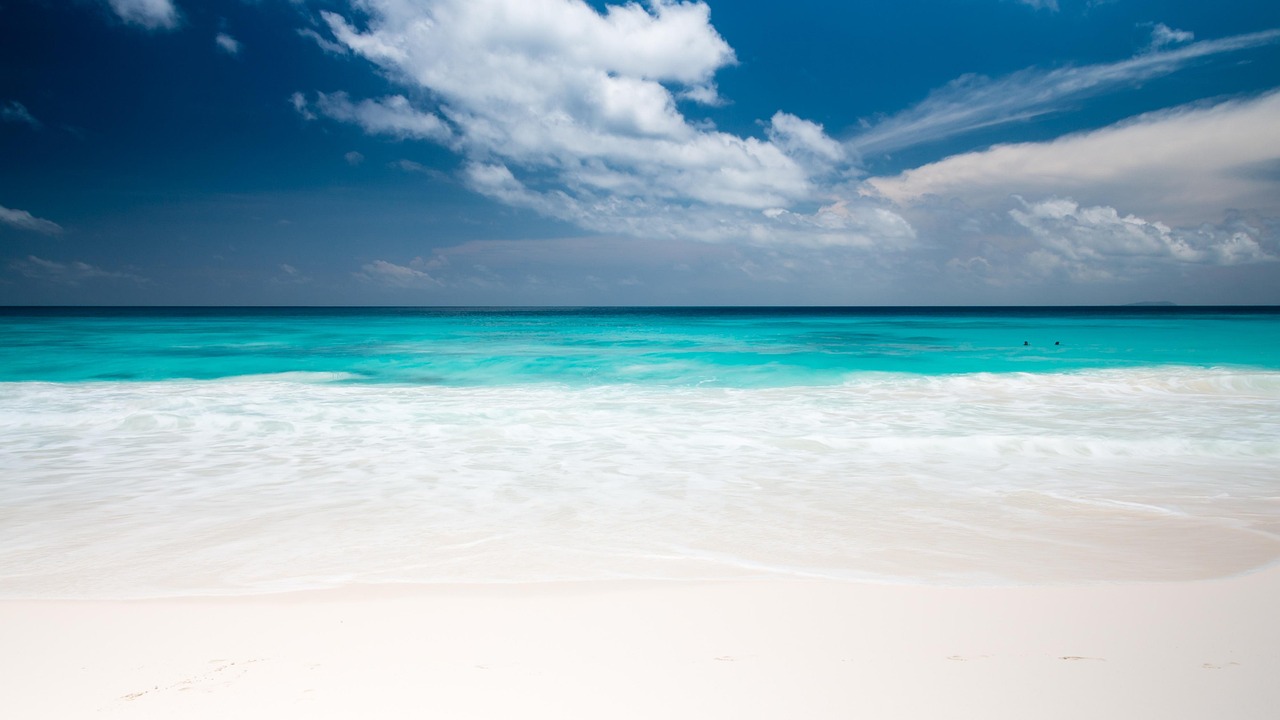Ein weisser Sandstrand mit tuerkisblauem Meer und weissen Wellen unter einem blauen Himmel mit Wolken.