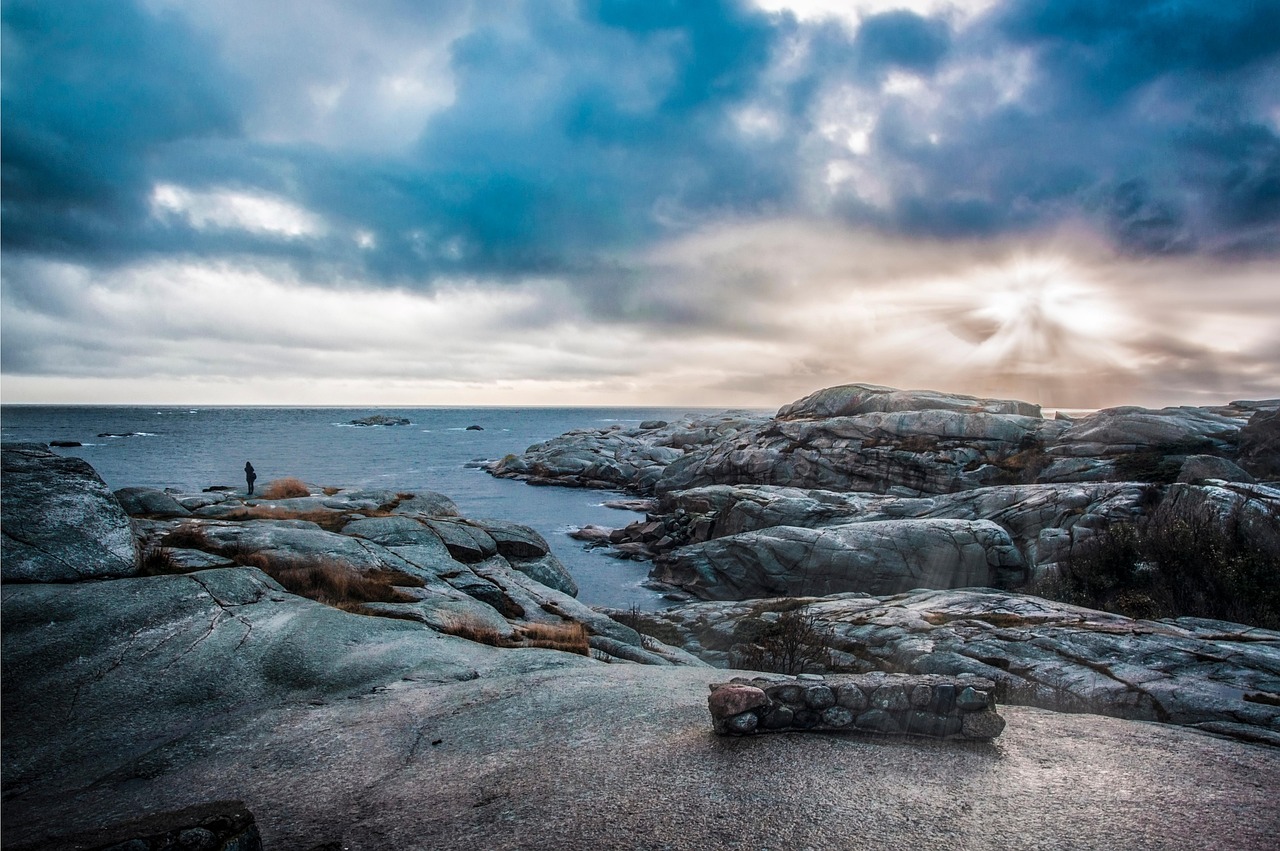 Eine Person steht auf einem felsigen Ufer und blickt auf das Meer unter einem dramatischen Himmel mit Wolken und Sonnenstrahlen.