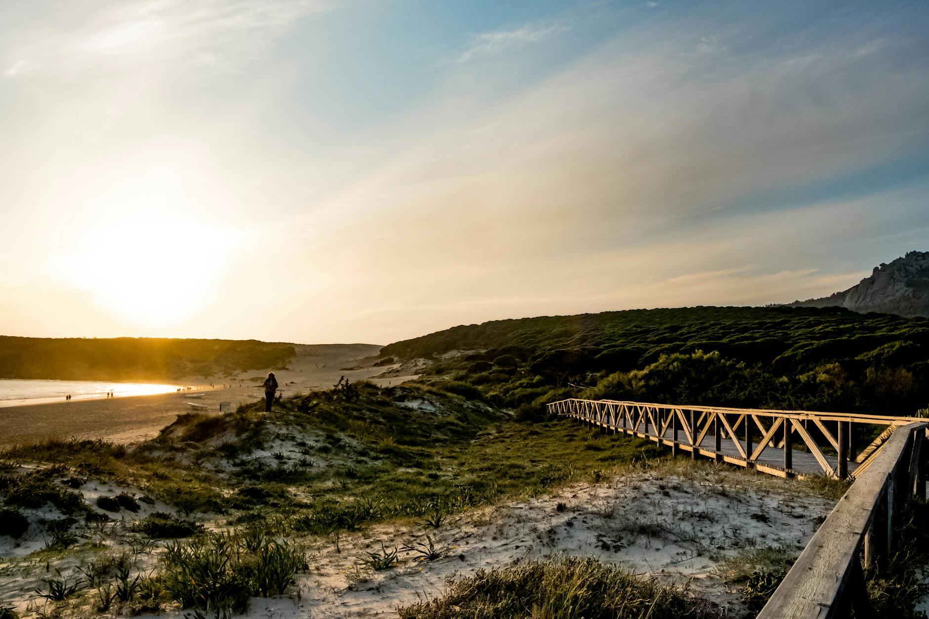 Ein Holzsteg fuehrt durch duenenbewachsene Landschaft zu einem Strand, an dem Menschen spazieren gehen, waehrend die Sonne untergeht.