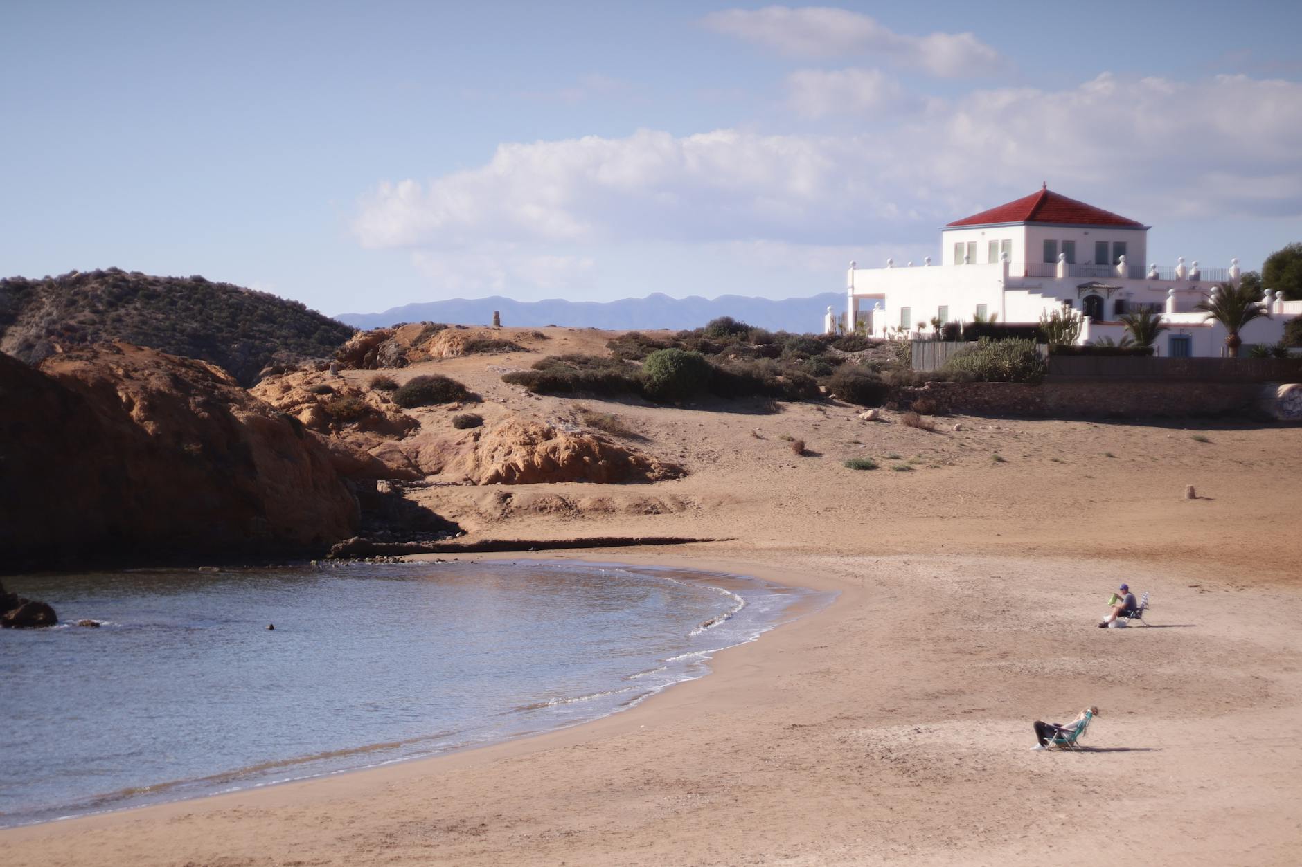 Ein Sandstrand mit einer Bucht, Felsen und einem weißen Haus mit rotem Dach im Hintergrund. Zwei Personen sitzen in Liegestühlen am Strand.