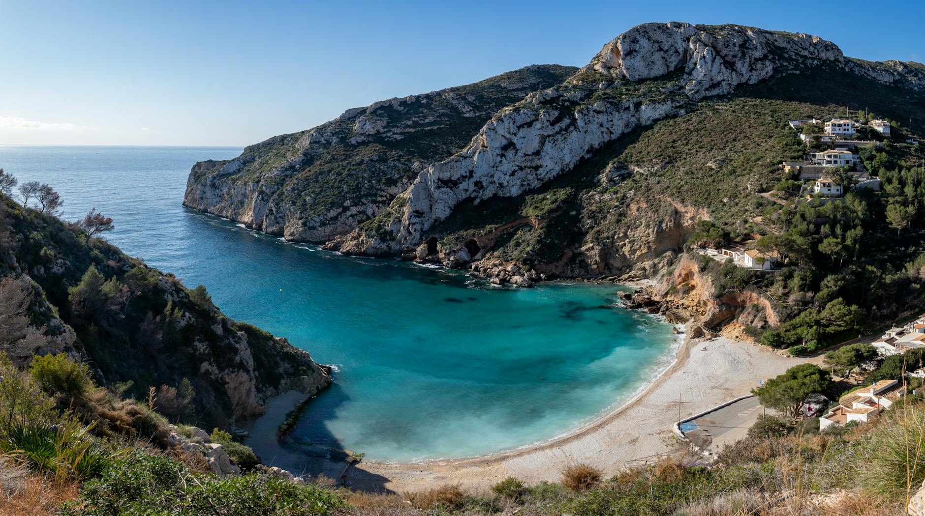 Eine malerische Bucht mit einem Kiesstrand und tuerkisem Wasser, umgeben von felsigen Hängen mit mediterraner Vegetation und einigen Ferienhäusern.