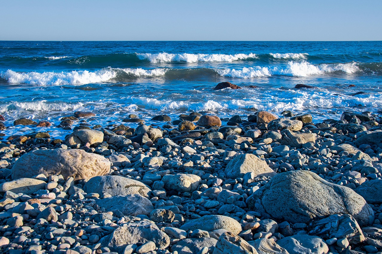 Ein Kiesstrand mit Wellen, die auf das Ufer treffen, unter einem klaren blauen Himmel.