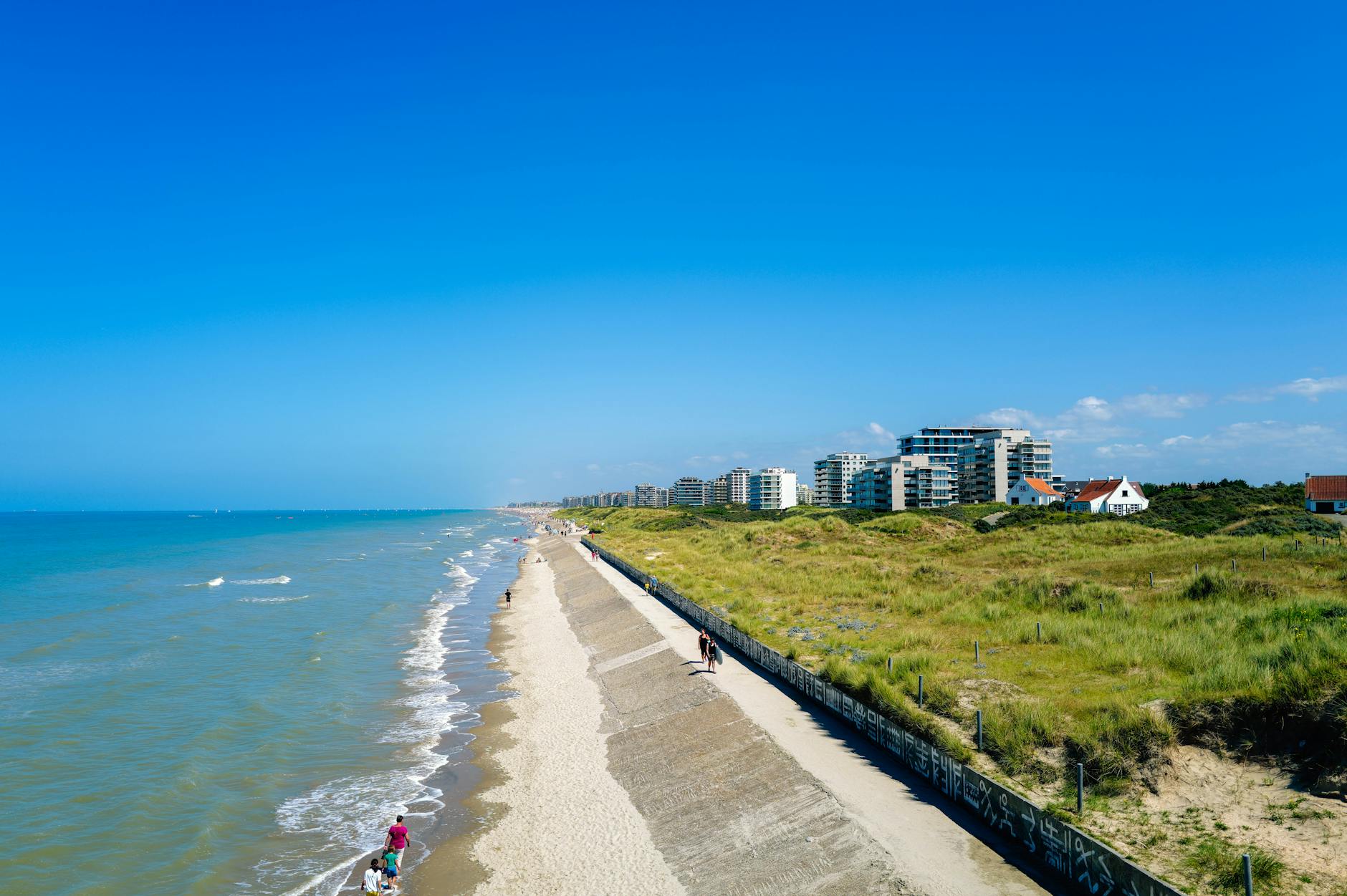 Ein breiter Sandstrand mit einer Promenade und dahinterliegenden Duenen und modernen Gebaeuden am Meer.
