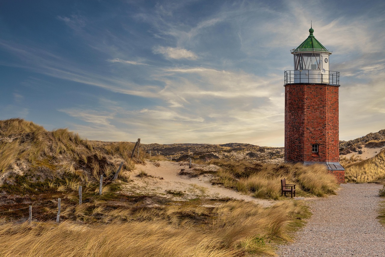 Ein roter Backsteinleuchtturm steht auf einer Düne neben einem Weg, umgeben von trockenem Gras und Büschen.