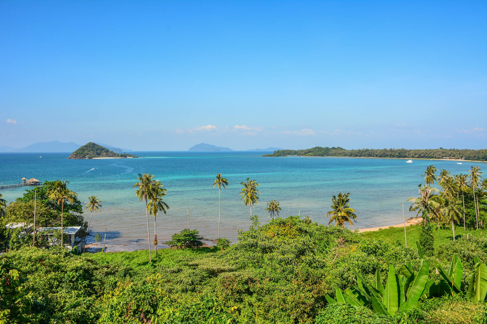 Blick auf einen tropischen Strand mit Palmen und türkisfarbenem Wasser, im Hintergrund eine kleine Insel und Berge.