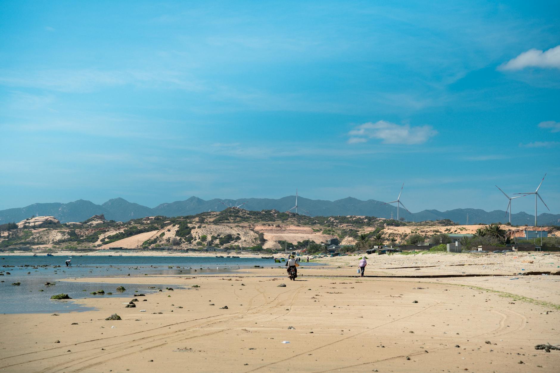 Ein Sandstrand mit dem Meer im Vordergrund und einer bergigen Landschaft mit Windrädern im Hintergrund.