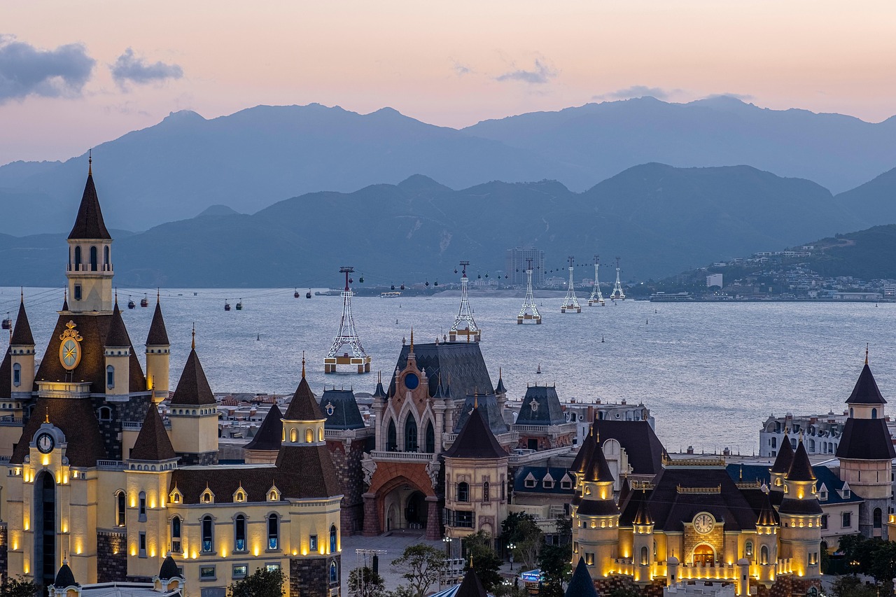 Eine Stadt mit schlossartigen Gebaeuden und Seilbahnen ueber dem Meer vor einer Bergkulisse bei Daemmerung.