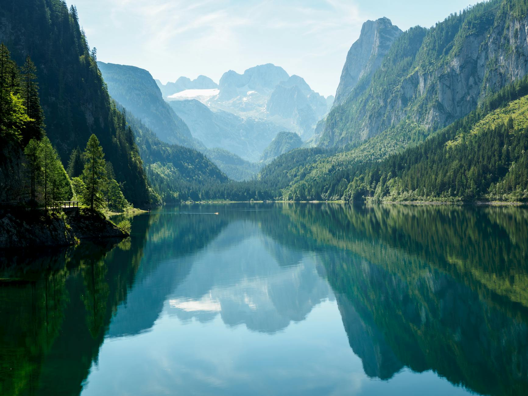 Ein ruhiger Bergsee mit Spiegelungen von bewaldeten Hängen und schneebedeckten Gipfeln im Hintergrund.
