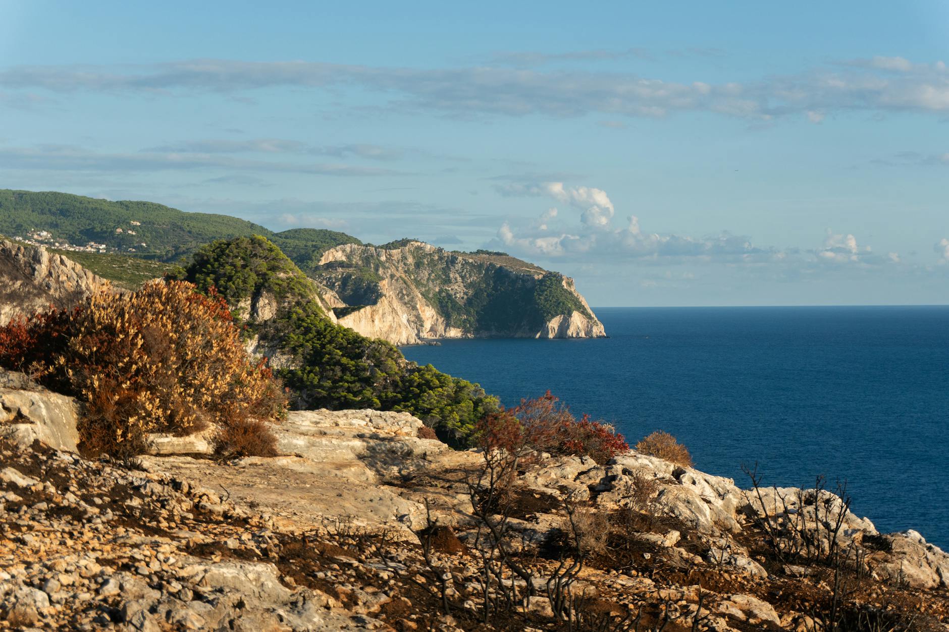 Eine felsige Küste mit verbrannter Vegetation im Vordergrund und grünen Hügeln im Hintergrund, die ins tiefblaue Meer abfallen.