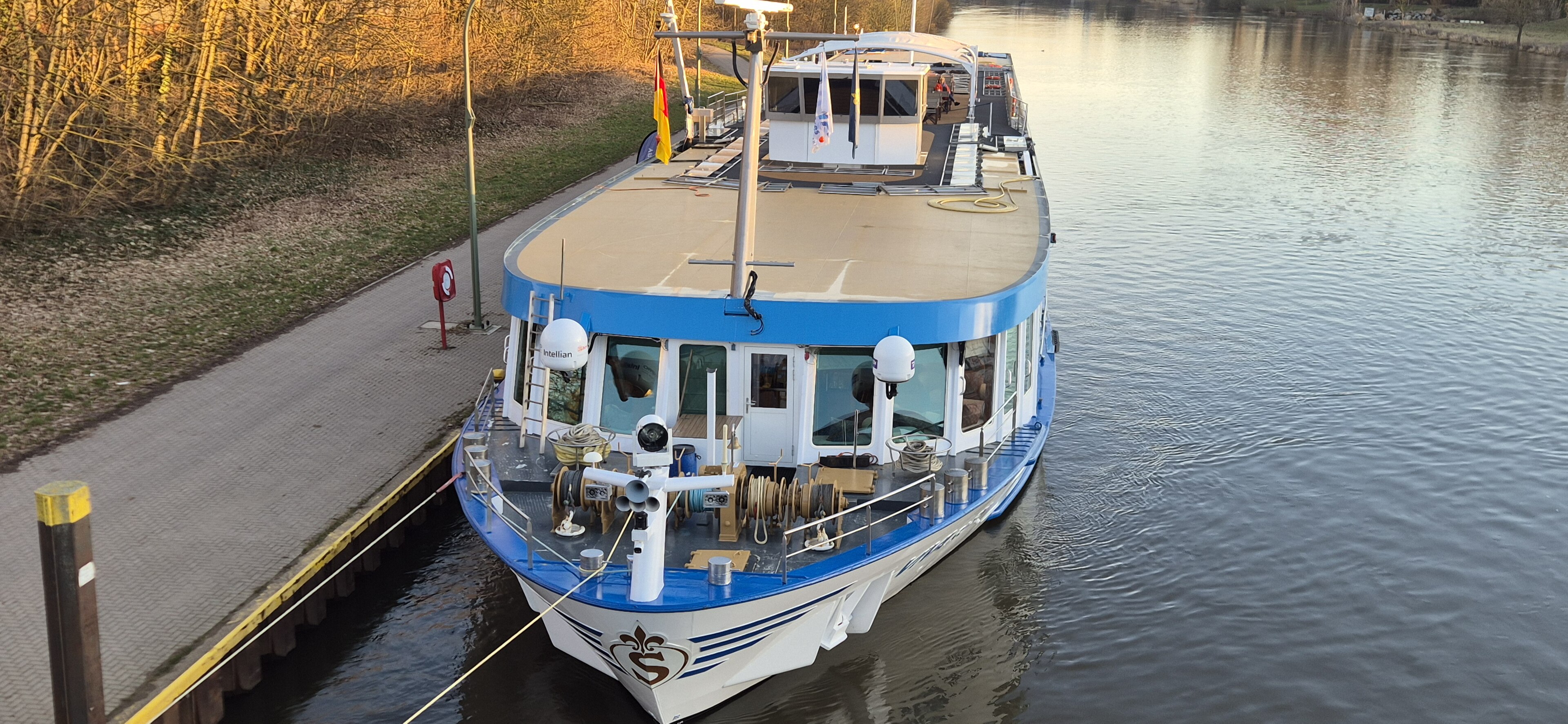 Ein blau-weißes Passagierschiff liegt an einer Anlegestelle an einem Fluss.